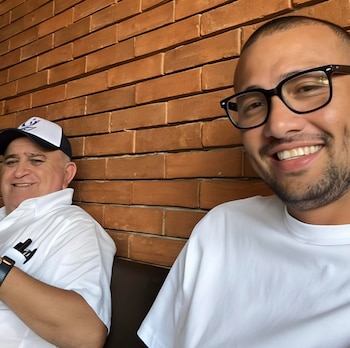 Dos hombres sonrientes sentados; uno mayor con gorra y camisa blanca, y uno joven con gafas y camiseta blanca. Pared de ladrillo de fondo