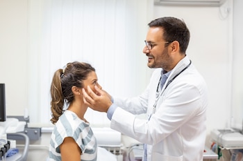 Un doctor con bata blanca y estetoscopio examina los ojos de una paciente sentada en un consultorio con equipo médico al fondo