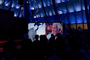 Personas sentadas en penumbra dentro de una sala con estructura metálica e iluminación azul mientras ven una pantalla proyectada