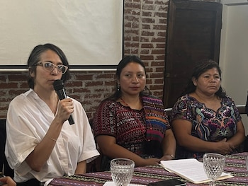 Tres mujeres sentadas en una mesa con mantel colorido. La del centro y derecha visten blusas tradicionales. La de la izquierda, con gafas, habla por micrófono
