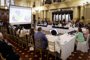 Reunión en un salón formal con columnas y vitrales. Personas sentadas en mesas largas frente a una pantalla proyectando un mapa de Guatemala con zonas de colores