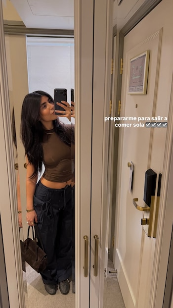 Mujer con cabello oscuro se toma selfie en espejo, viste top marrón y pantalones oscuros, sostiene bolso. Hay puerta con cartel de emergencia y texto