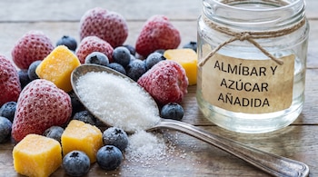 Frutillas, arándanos y trozos de mango congelados junto a una cuchara con azúcar y un frasco de vidrio con una etiqueta.
