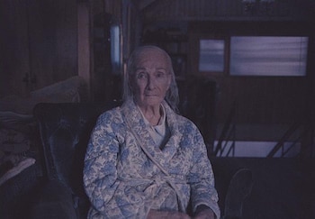 Una mujer de edad avanzada con bata de flores azul y blanca permanece sentada en una habitación en penumbra mirando al frente