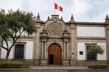 Fachada de piedra con portón de madera, un escudo de armas esculpido arriba, dos ventanas con rejas y la bandera de Perú en lo alto.
