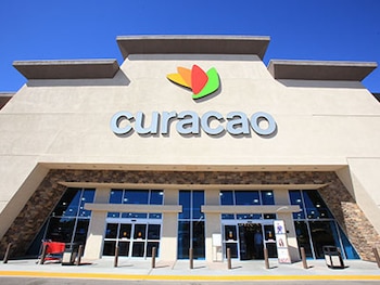 Fachada da loja de Curaçao com logotipo 'Curacao' e logotipo colorido. Portas de vidro e elementos de pedra podem ser vistos sob o céu azul