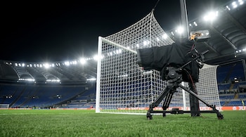 Cámara de televisión sobre un trípode colocada junto a la red de una portería en el césped de un estadio iluminado.