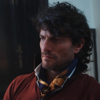 Un hombre de cabello oscuro y rizado, con barba, mira seriamente hacia la derecha. Viste una chaqueta roja y un pañuelo azul con diseño dorado