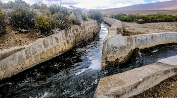Canal de agua construido con paredes de piedra y concreto en un terreno árido con vegetación baja y montañas.
