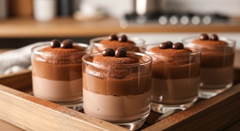 Seis vasos transparentes contienen un postre en capas de crema de chocolate y moka, espolvoreados con cacao y decorados con dos bolas de chocolate, sobre una bandeja de madera oscura.