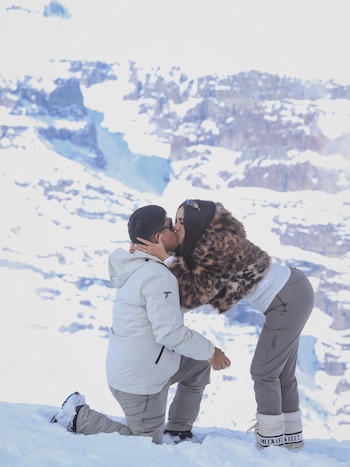Hombre arrodillado besa a mujer de pie en un paisaje de montaña nevado. Ambos visten ropa de invierno, con botas de nieve, bajo la luz del sol