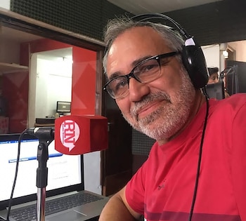 Hombre con gafas, barba y camiseta roja lleva auriculares negros frente a un micrófono de radio y una computadora portátil