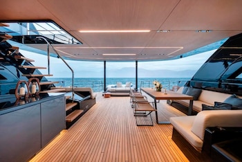 Cubierta de embarcación con suelo de teca, mesa de comedor, sillas, sofás y una escalera lateral con vista directa hacia el mar abierto