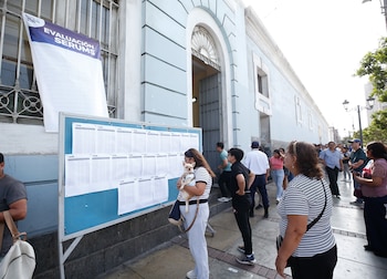 Grupo de personas mira un tablero informativo con numerosas hojas impresas. En la ventana cuelga un cartel azul y blanco que dice "EVALUACIÓN SERUMS"