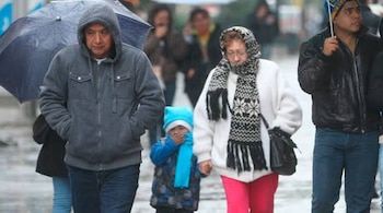 Personas con abrigos, bufandas, gorros y paraguas caminan por una calle húmeda