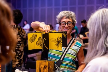 Mujer con cabello gris y gafas sujeta un libro plegable amarillo y negro con dibujos de insectos. Otros asistentes a la feria en segundo plano