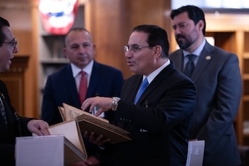 Un hombre de mediana edad con gafas y traje oscuro, que lleva una corbata azul, sostiene y hojea un libro antiguo de tapas marrones, con dos hombres borrosos detrás