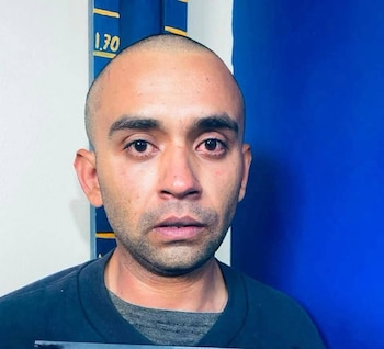 Primer plano de hombre con cabeza rapada, barba incipiente. Ojos enrojecidos. Viste ropa oscura. Fondo: pared blanca con medidor de altura y una sección azul
