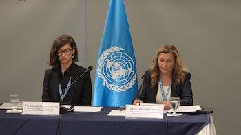 Dos mujeres están sentadas frente a micrófonos y letreros de identificación, con una bandera de las Naciones Unidas al fondo