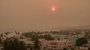 Vista aérea de una ciudad costera envuelta en calima o polvo. El sol rojizo es visible en el cielo brumoso sobre edificios, palmeras y el mar con barcos.