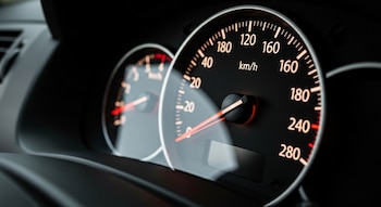 Primer plano del velocímetro de un automóvil con fondo oscuro. La aguja está en cero y los números de velocidad (km/h) están iluminados. Un segundo dial borroso es visible a la izquierda.