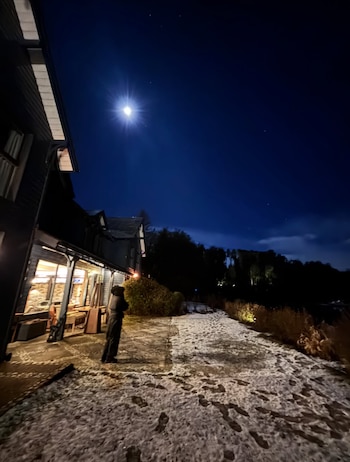 Una persona de espaldas está de pie sobre nieve en un sendero. Se observa un edificio oscuro con ventanas iluminadas y la luna brillando en el cielo nocturno