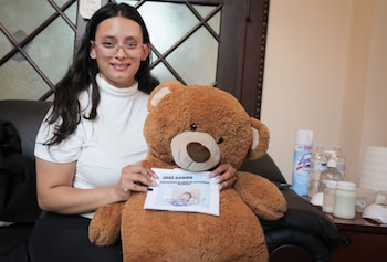 Una mujer sonriente con gafas y cabello oscuro sostiene un oso de peluche marrón grande que exhibe una tarjeta blanca con la foto de un bebé recién nacido