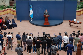 Un hombre en un podio junto a la bandera de Guatemala habla frente a un grupo de fotógrafos y periodistas