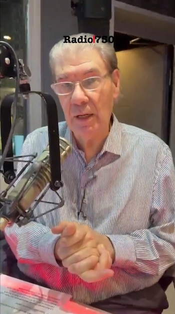 Un hombre de cabello gris y gafas, vestido con camisa a rayas, sentado en un estudio de radio frente a un micrófono. Sus manos están unidas.