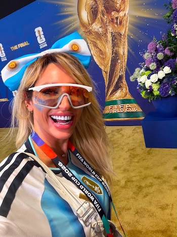 Mujer rubia sonriendo con gafas blancas, lazo con bandera argentina, camiseta de la selección, fondo azul con logo FIFA World Cup 2026 y réplica del trofeo