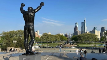 Estatua de bronce de Rocky con brazos levantados, de espaldas, frente a edificios de Filadelfia, árboles verdes y personas en el área del museo
