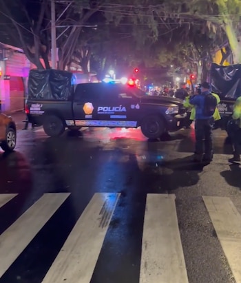 Una patrulla de policía con luces encendidas bloquea una calle de noche junto a varios agentes y un paso peatonal