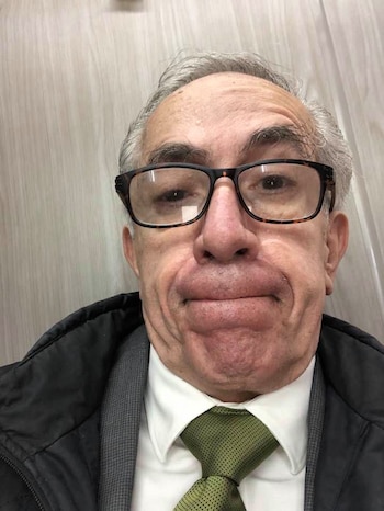 Primer plano de un hombre de mediana edad con cabello gris, gafas de montura oscura, camisa blanca, corbata verde y una chaqueta negra, mirando al frente