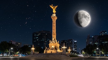 Monumento del Ángel de la Independencia iluminado de noche, con edificios al fondo y una luna en fase gibosa en un cielo estrellado.