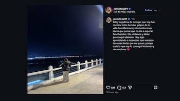 Una mujer de espaldas, con chaqueta oscura y pantalón claro, de pie en un muelle con barandales blancos, mirando el mar y una ciudad iluminada de noche
