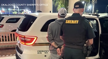 Un oficial del sheriff con chaleco amarillo "SHERIFF" pone a un hombre esposado, con gorra y sudadera gris, en la parte trasera de un coche patrulla blanco de noche