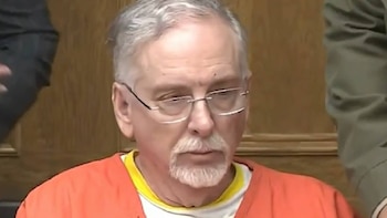 Primer plano de un hombre caucásico de edad avanzada, James M. Heaps, con pelo canoso, barba, bigote y gafas, vistiendo un uniforme naranja