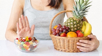Persona rechaza un cuenco de dulces con la mano y sostiene una cesta con piña, uvas, plátanos, naranjas y fresas en una ilustración de acuarela.