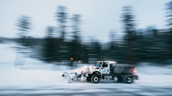 Un camión quitanieves blanco con una pala frontal se desplaza hacia la derecha sobre una carretera cubierta de nieve, con árboles borrosos al fondo