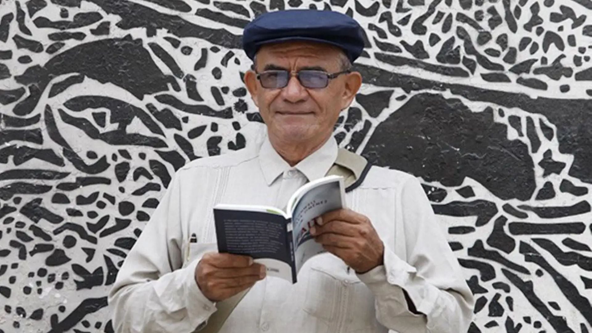 Cronwell Jara Jiménez, el escritor peruano, lee un libro con la cubierta parcialmente visible, de pie frente a un mural con diseño abstracto blanco y negro.