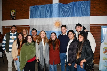 Un grupo de diez personas, cinco hombres y cinco mujeres, posan de pie frente a una bandera argentina y un retrato de Eva Perón en una pared de ladrillos