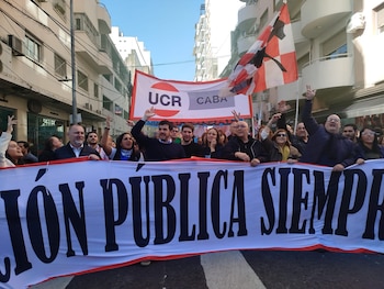 Multitud de personas marchando por una calle, sosteniendo una pancarta blanca con el texto "EDUCACIÓN PÚBLICA SIEMPRE" y un cartel rojo y blanco de UCR CABA