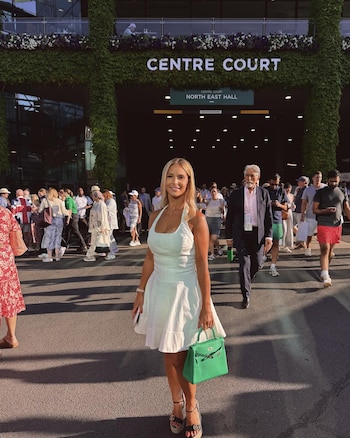 Laura Hilven, de pie, con vestido blanco y bolso verde, sonríe frente a la entrada del Centre Court de Wimbledon, rodeada de multitudes