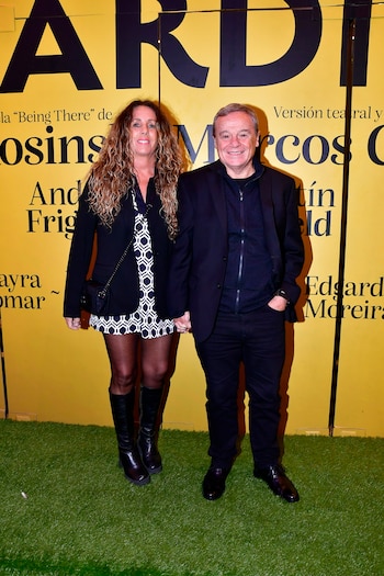 Dos personas, una mujer con cabello rizado y un hombre, se toman de la mano y sonríen frente a un fondo amarillo con letras negras y césped verde artificial