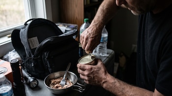 Hombre abre una lata de conserva de metal, un bol con pescado, un tenedor, una mochila negra, dos linternas, dos botellas de agua