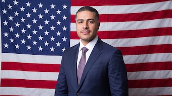 Omar García Harfuch vestido con traje azul y corbata posa de frente ante la bandera de Estados Unidos.