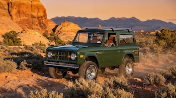 Un Ford Bronco verde oscuro de época con parrilla cromada y neumáticos todoterreno sobre un camino de tierra rocoso, con montañas y rocas al fondo al atardecer.