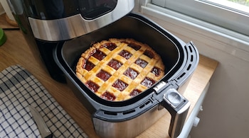 Una pastafrola de membrillo dentro de la canasta de una freidora de aire negra y plateada sobre una encimera de madera junto a una ventana.