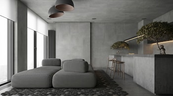 Interior moderno con sofá modular gris, alfombra de espiral y barra de hormigón. Ventanas grandes, luces colgantes y plantas decoran el espacio.