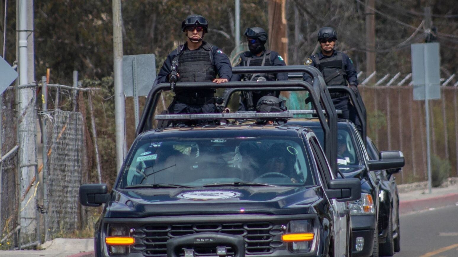 Elementos de seguridad, vestidos con uniforme táctico oscuro, se desplazan en vehículos patrulla por una vía urbana.